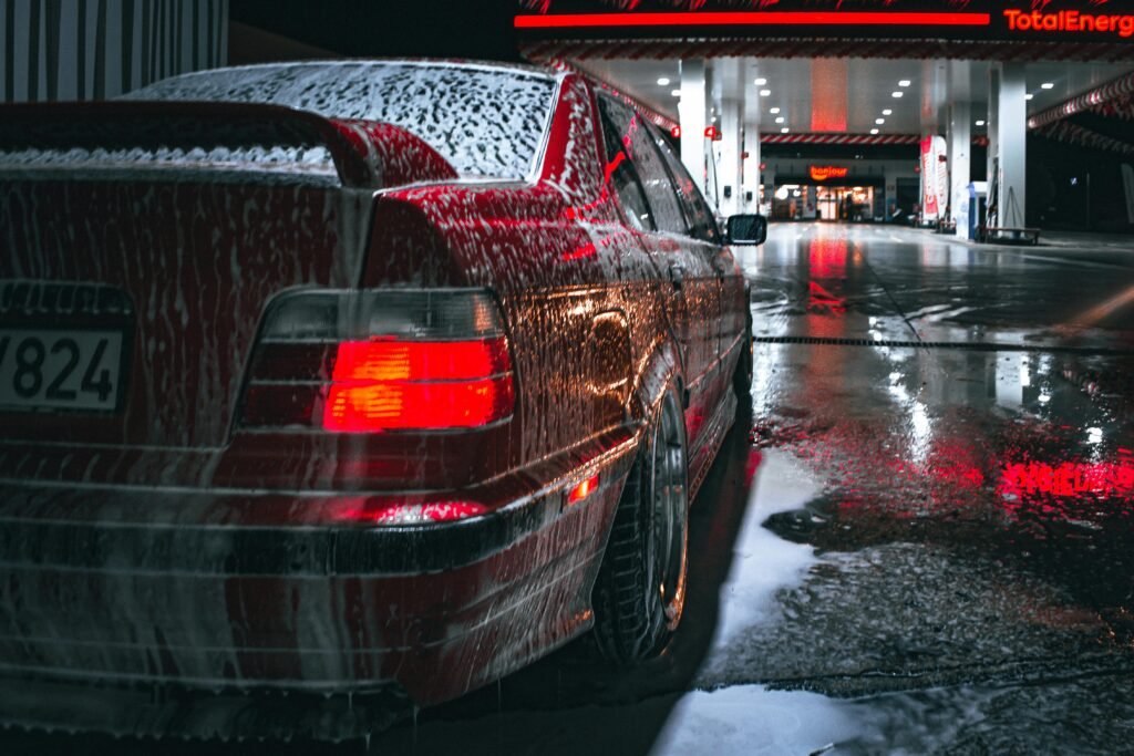 Red car covered in soap at night near a gas station with bright lights reflecting on wet pavement.